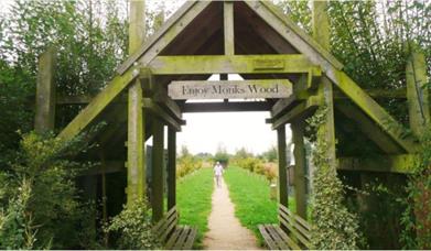 A wooden hut structure surrounded by trees with a sign that reads 'Enjoy Monks Wood', with benches and a path stretching through the centre.