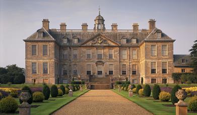 A driveway leading to a large manor house, with small hedges lining the way to the entrance.