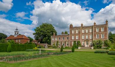 A symmetrical historic house towers proudly over an immaculate lawn, bordered by hedges
