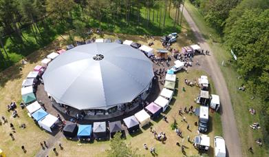 A large circle big top surrounded by market stalls in a field with trees
